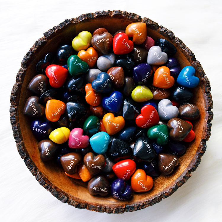 Colorful hand-carved soapstone hearts from Kenya, featuring intricate art carvings and inspirational words, arranged in a bowl. Each heart showcases vibrant hues and unique designs.